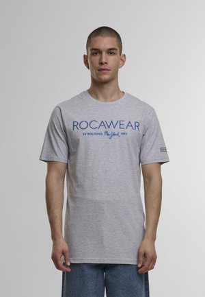 Pelēka kokvilnas T-kreklu ar zilo ROCAWEAR logo un dibināšanas datumu. Īsas piedurknes, apaļa kakla izgriezums, taisna apakšmala un standarta izmērs.
