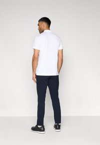 Άσπρο κοντομάνικο πόλο πουκάμισο, ναυτικό μπλε παντελόνι, μαύρα παπούτσια. Το πόλο διαθέτει γιακά και πλαϊνά σκισίματα. Προβολή από πίσω, ουδέτερο φόντο.