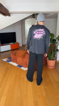 Hosszú ujjú szürke ing színes szöveggel és grafikával a hátoldalán, fekete bőszárú nadrággal párosítva. Laza környezet növénnyel.