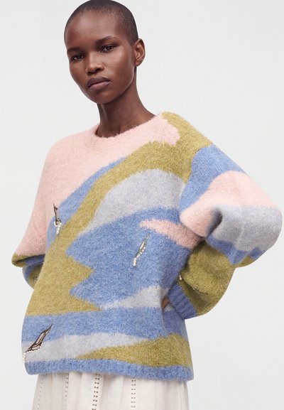 Femme portant un pull doux pastel avec des motifs abstraits bleu, vert, rose et gris et une petite broderie d'oiseau sur un fond clair uni.