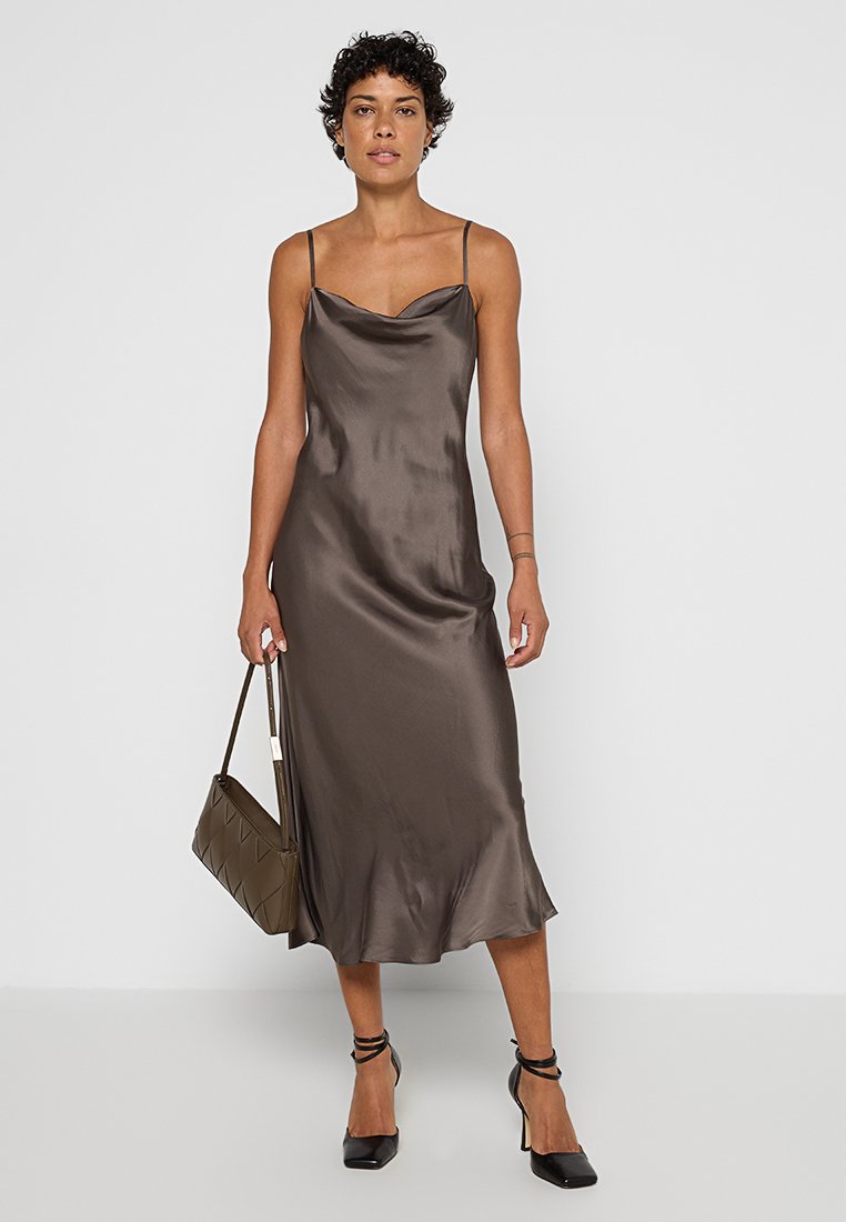Mujer que lleva un vestido de satén marrón con tirantes finos, tacones negros atados al tobillo y sostiene un bolso marrón frente a un fondo liso.