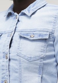 Light blue denim jacket with a collar, front button closure, and two chest pockets. Features silver button hardware and a slightly faded texture.