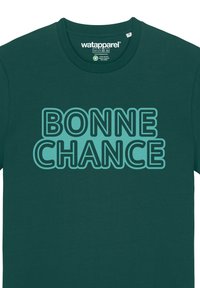 T-shirt vert foncé affichant le texte "BONNE CHANCE" en bleu clair avec des lettres bold et en contour. Fabriqué dans un matériau lisse et doux.