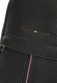 Μαύρο δερμάτινο σακίδιο με υφή στην επιφάνεια, διαθέτει οριζόντια φλάπα και διακοσμητική ρίγα από κόκκινες, λευκές και μπλε ραφές. Εμφανίζεται το λογότυπο.