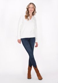 Weißer V-Ausschnitt-Pullover mit Zopfstrick-Ärmeln, kombiniert mit dunkelblauen Jeans und braunen Wildlederschnürstiefeln mit spitzem Oberteil. Glatte Textur.