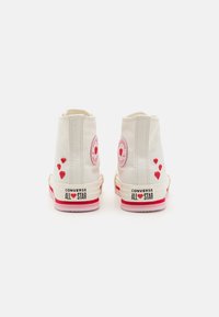 Plátené tenisky so zvýšeným členkom v krémovej farbe s červenými srdcovými akcentmi, značkou Converse a ružovou gumovou podrážkou. Sú zdobené vyšívanými detailmi.