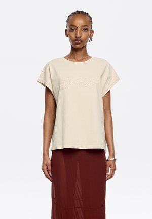 T-shirt beige a maniche corte con scritta ricamata "Bimba y Lola", dalla vestibilità morbida e dalla texture liscia, abbinato a una gonna rossa semi-trasparente.