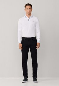 Camiseta polo blanca de manga larga con cuello a rayas azul marino, combinada con vaqueros negros y zapatillas oscuras, de pie frente a un fondo neutro.