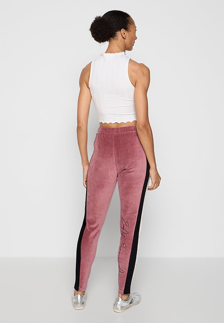 Jasnoszary krótki top z falbaniastym dekoltem, zestawiony z różowymi dresami z weluru, które mają czarne boczne paski i charakterystyczny detal.