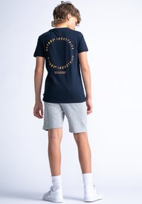 Navy katoenen T-shirt met een ronde oranje grafiek op de achterkant, gecombineerd met grijze shorts. Model staat met de handen langs het lichaam.