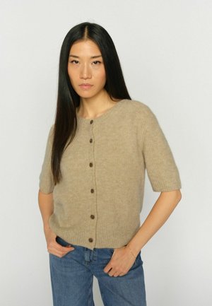 Frau mit langen schwarzen Haaren, die einen beigen, kurzärmeligen, geknöpften Pullover und blaue Jeans trägt, steht mit den Händen in den Taschen vor einem schlichten Hintergrund.