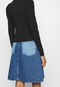 Schwarzes Langarmoberteil mit gepufften Schultern, kombiniert mit einem zweifarbigen blauen Jeansrock mit hellen und dunklen Waschungen sowie hinteren Taschen.