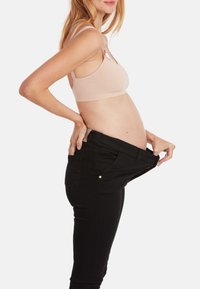 Un soutien-gorge de maternité beige et un jean stretch noir tenus par la taille, montrant un ventre de femme enceinte. Tissu doux et design ajusté visibles.