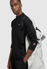 Fekete kapucnis pulóvert viselő férfi, rajta a "KHUJO" logóval és az "01" számmal az ujján, fehér pufferek zakót tart a válla fölött, egy sima szürke háttér előtt.