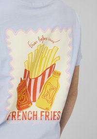 Frites dans un contenant rouge et blanc rayé avec des bouteilles de ketchup et de mayonnaise, texte « fries before cries » sur un t-shirt bleu clair.