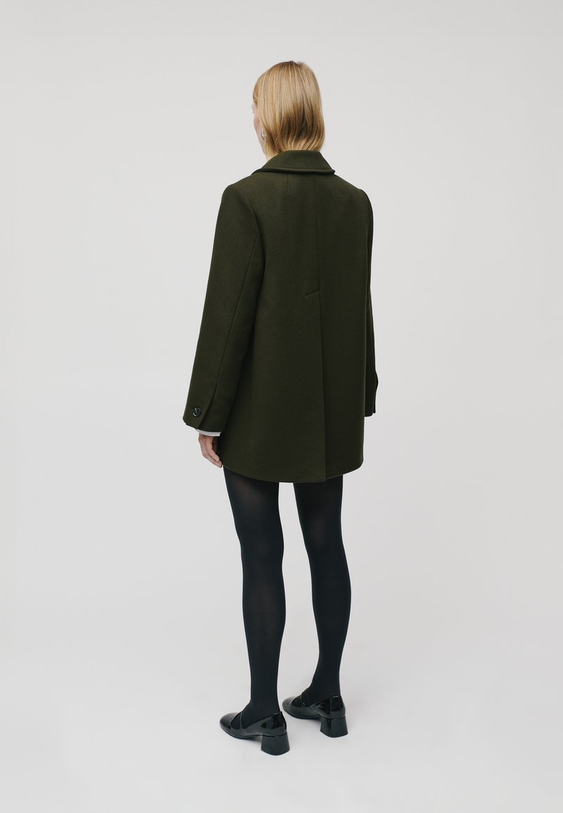 Manteau ajusté vert foncé avec un col, fente simple à l'arrière et manches boutonnées. Porté par-dessus des collants noirs et des chaussures noires brillantes.