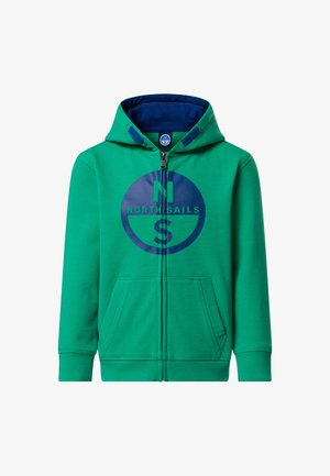 Garçon aux cheveux bruns portant un sweat à capuche vert zippé et un pantalon bleu marine, tenant une veste bleu marine dans sa main gauche, debout sur un fond uni.