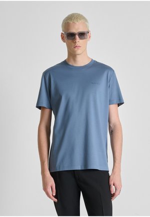 Giovane uomo con capelli biondi corti che indossa occhiali da sole scuri, una maglietta blu e pantaloni neri, in piedi davanti a uno sfondo grigio uniforme.