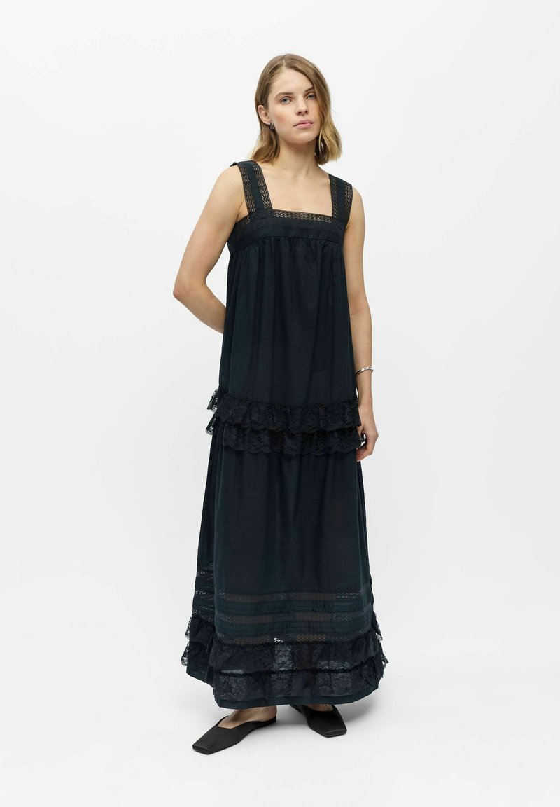 Femme portant une robe maxi noire sans manches avec des volants en dentelle, debout avec une main derrière le dos sur un fond blanc uni.