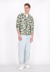 Giacca verde con zip decorata con un motivo di fenicotteri e foglie di palma; abbinata a pantaloni cargo azzurri e sneakers bianche.