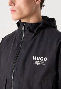 Musta huppari, jossa on vetoketjullinen etuosa, jossa on näkyvä valkoinen logo "HUGO" ja teksti. Valmistettu sileästä, vedenkestävästä materiaalista.
