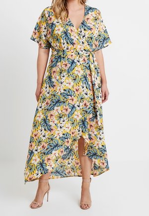 Kvinde iført en gul blomsterprintet slå-om-kjole med blå og pink blomster, korte ærmer, frontslids og nøgne høje hæle sandaler.