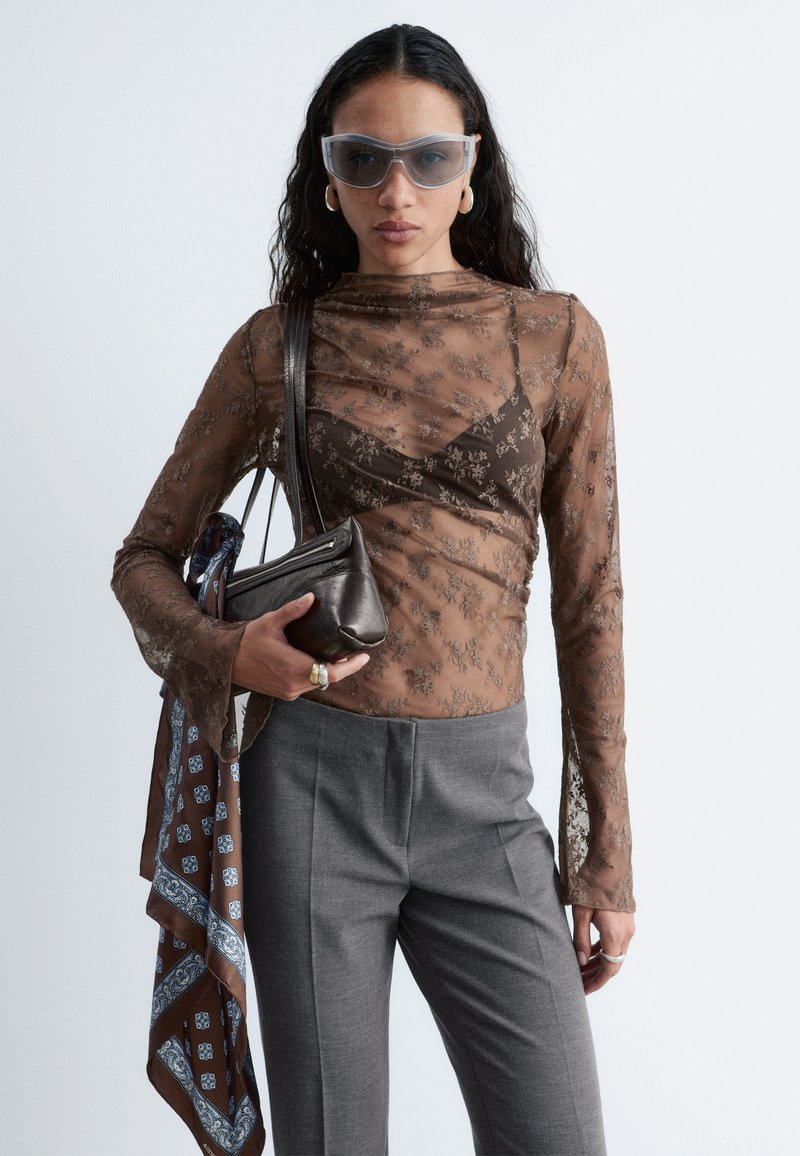Femme portant un haut marron fleuri transparent sur un soutien-gorge noir, un pantalon gris, des lunettes de soleil transparentes, tenant un sac à main noir avec une écharpe marron à motifs.