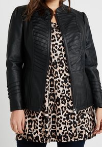 Zwarte faux leren jas met geribbelde details op de schouders en aan de voorkant, met een getailleerde silhouette en ritssluiting, gelaagd over een top met luipaardprint.