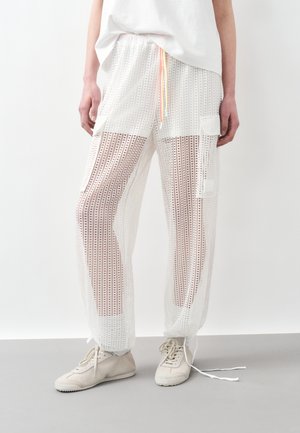 Person trägt weiße Mesh-Cargohosen mit Kordelzugbund, weiße Sneakers und ein weißes Hemd vor einem schlichten hellen Hintergrund.