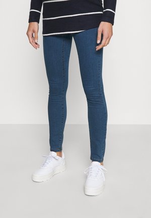 Persoon die donkerblauwe skinny jeans, witte sneakers en een zwarte trui met witte horizontale strepen draagt, staand op een vlakke vloer.