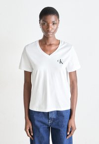 Tricou alb din bumbac cu decolteu în V, mâneci scurte și un mic logo negru "CK" pe partea stângă a pieptului. Textură netedă, croială relaxată.