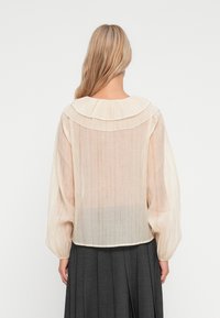Lett, gjennomsiktig bluse i beige med et lagdelt, rundt krage, puffede ermer og en plisserte bakhem. Teksturert stoffdetalj synlig.
