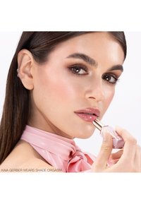 Femme avec un maquillage des yeux brillant et un foulard en satin rose appliquant un rouge à lèvres rose clair en regardant vers la caméra, vue rapprochée.