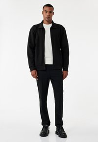 Sort zip-jakke med krave, båret over en hvid sweater. Slanke sorte bukser og sorte sko fuldender outfittet. Glat tekstur, minimalistisk design.
