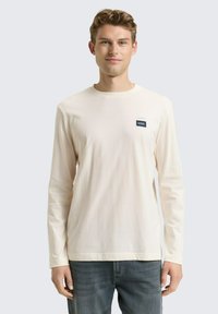 Kremowa koszulka z długim rękawem wykonana z miękkiej bawełny, z okrągłym dekoltem i małą granatową metką po lewej stronie klatki piersiowej. Noszona z niebieskimi dżinsami.