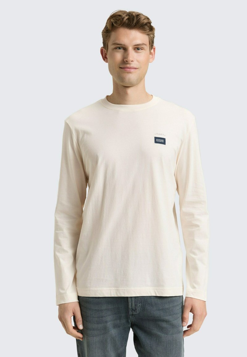 Kremowa koszulka z długim rękawem wykonana z miękkiej bawełny, z okrągłym dekoltem i małą granatową metką po lewej stronie klatki piersiowej. Noszona z niebieskimi dżinsami.