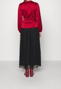Blouse en satin rouge avec de longues manches bouffantes et une taille cintrée, associée à une jupe maxi en tulle à pois noirs et des bottes à talons chunky rouges.