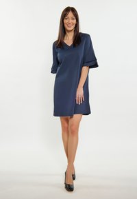 Marineblaues Leinenkleid mit V-Ausschnitt und kurzen, gerafften Ärmeln. Knielanges Design, glatte Textur, kombiniert mit schwarzen High Heels.