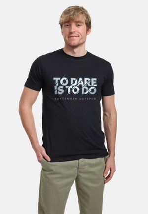 Giovane uomo che indossa una T-shirt nera con testo bianco "To Dare Is To Do" e "Tottenham Hotspur", abbinata a pantaloni verde oliva chiaro.