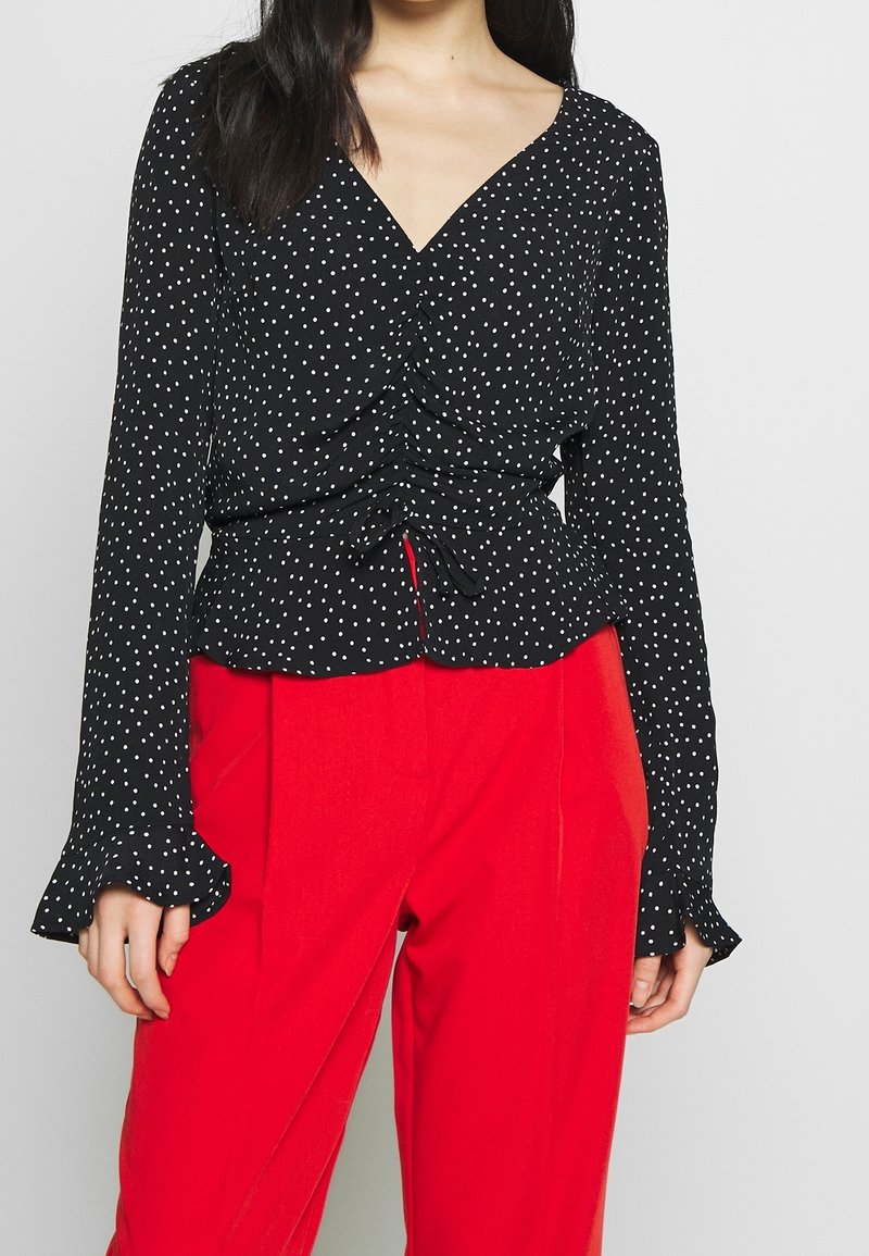 Blusa nera a pois con design avvolgente, vita arricciata, maniche lunghe con polsini svasati, abbinata a pantaloni rossi su misura.