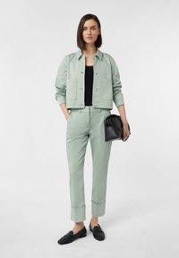 Jasnozielona krótka kurtka z zapięciem na guziki i dwoma przednimi kieszeniami, w połączeniu z dopasowanymi, zwężającymi się spodniami oraz czarnymi mokasynami, trzymająca czarną torbę.