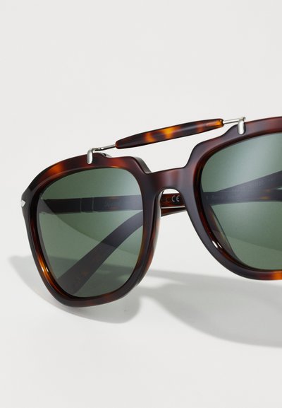 Καφέ καβουροειδή γυαλιά ηλίου με υπερμεγέθη τετράγωνα φάσματα, σκούρους πράσινους φακούς και ασημένιες μεταλλικές λεπτομέρειες στη γέφυρα και τους ναούς.