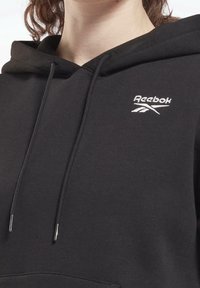 Felpa nera Reebok con tasca frontale, cappuccio regolabile con stringhe e logo ricamato. Tessuto morbido con una texture liscia e una vestibilità comoda.
