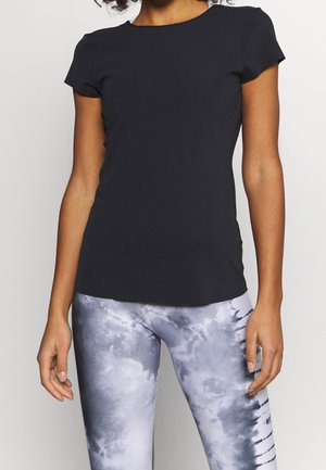 Vrouw met een nauwsluitend zwart kortemouwshirt en grijze tie-dye leggings, staand tegen een eenvoudige lichte achtergrond.