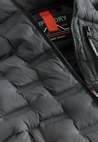 Puffet sort jakke med quiltet design, lynlås-lommer, orange indvendig mærke og logo-detalje. Fremstillet af et glat, let materiale.