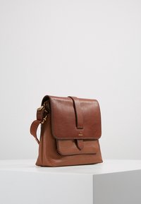 Brązowa skórzana torba na ramię z klapką i regulowanym paskiem, wyeksponowana na białym piedestale na jednolitym tle.