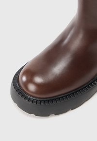 Botín de cuero marrón con un acabado suave y brillante. Presenta una suela de goma negra gruesa con detalles acanalados y una forma de puntera redondeada.