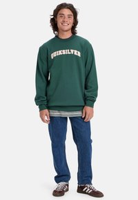 Zelený svetrový sveter s krúžkovým golierom a krémovým logom "Quiksilver". V kombinácii s pruhovanou košeľou pod ním a modrými džínsami. Hnedé tenisky.