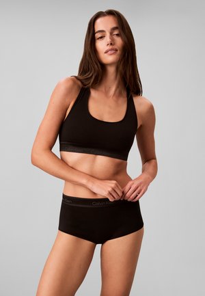 Femme portant un soutien-gorge de sport noir Calvin Klein et un sous-vêtement noir assorti sur un fond gris uni.