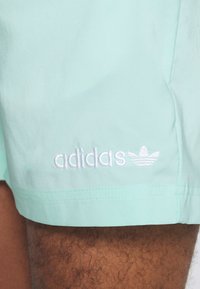 MIntgrønne shorts med en glat tekstur, med hvidt broderet "adidas"-tekst og logo nederst på kanten.